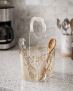 Clear acrylic spoon holder and cutlery caddy with gold accent patterns and a heart-shaped handle on a kitchen countertop holding utensils.