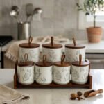 A matching 7-piece ceramic kitchen storage jar set with white and green leaf patterns, wooden lids featuring pull loops, and a tiered wooden organizer stand on a white kitchen countertop.