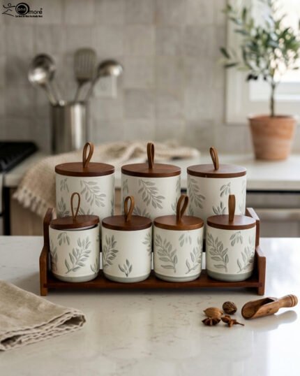 A matching 7-piece ceramic kitchen storage jar set with white and green leaf patterns, wooden lids featuring pull loops, and a tiered wooden organizer stand on a white kitchen countertop.