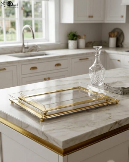 Two stacked luxury clear acrylic serving trays with polished gold trim and feet displayed on a white marble kitchen island.
