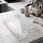 Set of clear acrylic serving trays with built-in handles and textured bases displayed on a marble kitchen counter next to an espresso machine and ceramic cups.