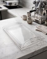 Set of clear acrylic serving trays with built-in handles and textured bases displayed on a marble kitchen counter next to an espresso machine and ceramic cups.