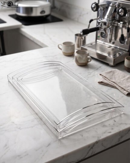 Set of clear acrylic serving trays with built-in handles and textured bases displayed on a marble kitchen counter next to an espresso machine and ceramic cups.