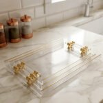 A nested set of clear ribbed acrylic serving trays with elegant gold handles and accents, displayed on a modern marble kitchen countertop next to glass spice jars.