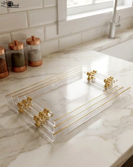 A nested set of clear ribbed acrylic serving trays with elegant gold handles and accents, displayed on a modern marble kitchen countertop next to glass spice jars.
