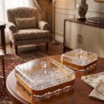 Two square crystal dry fruit dishes with gold trim bases and glass lids displayed on a wooden table in a luxury living room setting.