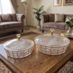 Two transparent crystal dry fruit serving dishes with partitioned interiors, gold geometric patterns, and crystal-topped lids placed on a wooden table.