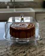 Clear acrylic cake stand with a domed lid and crystal knob displaying a frosted caramel-drizzled cake on a kitchen countertop.