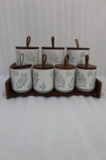 A matching 7-piece ceramic kitchen storage jar set with white and green leaf patterns, wooden lids featuring pull loops, and a tiered wooden organizer stand on a white kitchen countertop.