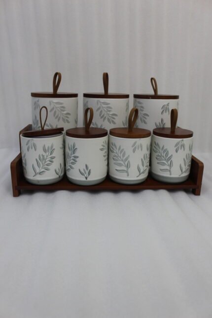 A matching 7-piece ceramic kitchen storage jar set with white and green leaf patterns, wooden lids featuring pull loops, and a tiered wooden organizer stand on a white kitchen countertop.