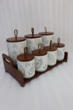 A matching 7-piece ceramic kitchen storage jar set with white and green leaf patterns, wooden lids featuring pull loops, and a tiered wooden organizer stand on a white kitchen countertop.