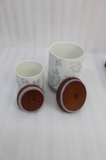 A matching 7-piece ceramic kitchen storage jar set with white and green leaf patterns, wooden lids featuring pull loops, and a tiered wooden organizer stand on a white kitchen countertop.