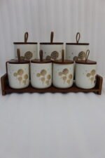 White ceramic kitchen storage jars with gold dandelion print, wooden lids with loop handles, and a wooden rack on a kitchen counter.