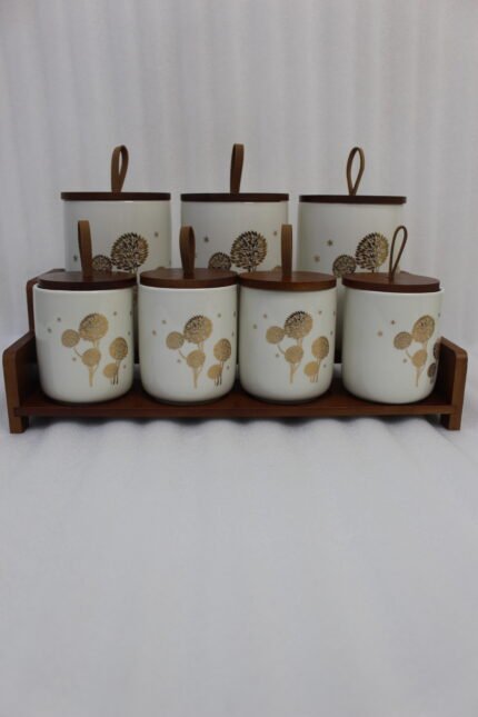 White ceramic kitchen storage jars with gold dandelion print, wooden lids with loop handles, and a wooden rack on a kitchen counter.