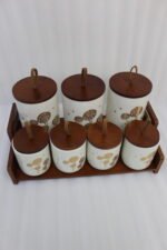 White ceramic kitchen storage jars with gold dandelion print, wooden lids with loop handles, and a wooden rack on a kitchen counter.