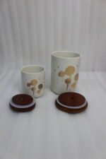 White ceramic kitchen storage jars with gold dandelion print, wooden lids with loop handles, and a wooden rack on a kitchen counter.
