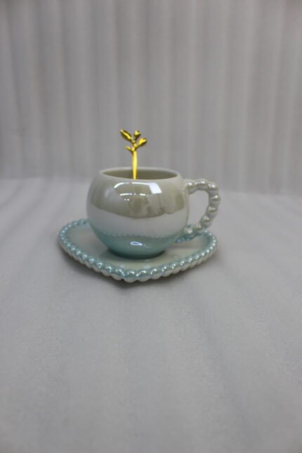 Elegant pastel ceramic cup and saucer set with a matching gold spoon and beaded handle on a marble kitchen countertop.