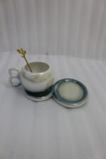 A collection of multicolored ceramic coffee cups and saucers with gold leaf spoons and double-loop handles arranged on a marble kitchen counter.