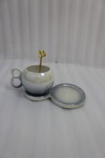 A collection of multicolored ceramic coffee cups and saucers with gold leaf spoons and double-loop handles arranged on a marble kitchen counter.