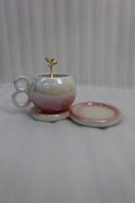 A collection of multicolored ceramic coffee cups and saucers with gold leaf spoons and double-loop handles arranged on a marble kitchen counter.