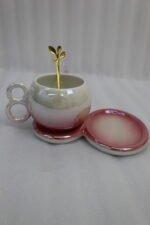 A collection of multicolored ceramic coffee cups and saucers with gold leaf spoons and double-loop handles arranged on a marble kitchen counter.