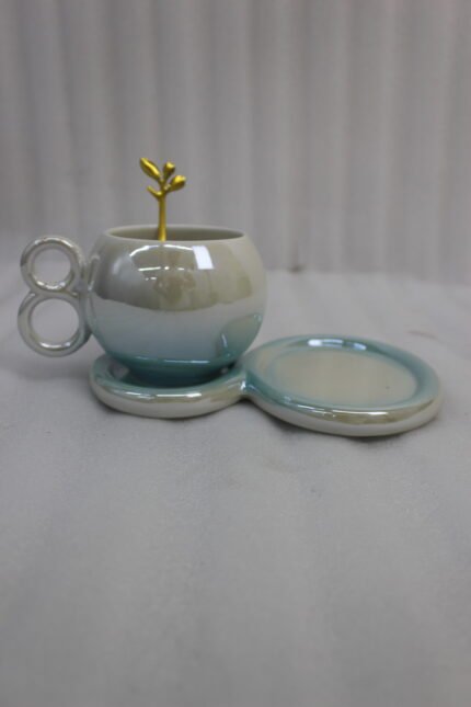 A collection of multicolored ceramic coffee cups and saucers with gold leaf spoons and double-loop handles arranged on a marble kitchen counter.
