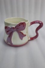 Four pastel ceramic coffee mugs with cute 3D bows and heart-shaped handles lined up on a kitchen counter.