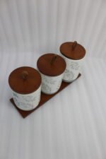Three white ceramic kitchen canisters with gray leaf patterns, wooden lids with loop handles, and a wooden tray displayed on a white kitchen countertop.