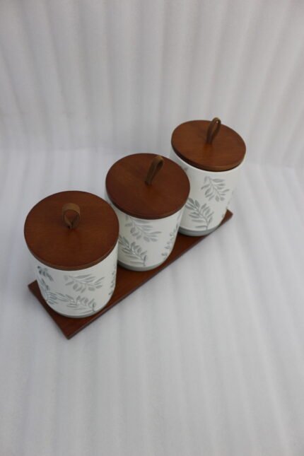 Three white ceramic kitchen canisters with gray leaf patterns, wooden lids with loop handles, and a wooden tray displayed on a white kitchen countertop.