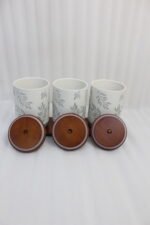 Three white ceramic kitchen canisters with gray leaf patterns, wooden lids with loop handles, and a wooden tray displayed on a white kitchen countertop.