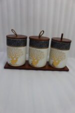 Three white and speckled black kitchen storage canisters featuring a gold deer and tree motif, fitted with wooden lids with loop handles and arranged on a matching wooden tray on a kitchen counter.