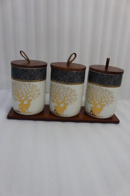 Three white and speckled black kitchen storage canisters featuring a gold deer and tree motif, fitted with wooden lids with loop handles and arranged on a matching wooden tray on a kitchen counter.