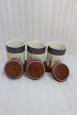 Three white and speckled black kitchen storage canisters featuring a gold deer and tree motif, fitted with wooden lids with loop handles and arranged on a matching wooden tray on a kitchen counter.