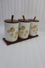 Three cream-colored ceramic kitchen storage jars featuring gold floral patterns, wooden lids with leather straps, and a matching wooden base tray sitting on a marble countertop.