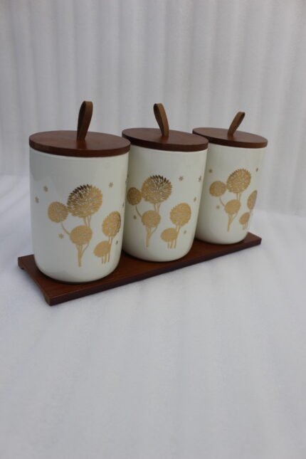 Three cream-colored ceramic kitchen storage jars featuring gold floral patterns, wooden lids with leather straps, and a matching wooden base tray sitting on a marble countertop.