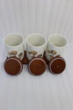 Three cream-colored ceramic kitchen storage jars featuring gold floral patterns, wooden lids with leather straps, and a matching wooden base tray sitting on a marble countertop.