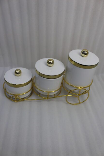 A set of three white ceramic jars with gold lids and spherical handles, elegantly arranged in a decorative gold metal cart stand against a white background.