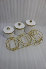 A set of three white ceramic jars with gold lids and spherical handles, elegantly arranged in a decorative gold metal cart stand against a white background.