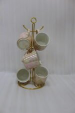 A set of 6 iridescent ceramic tea and coffee mugs hanging on a gold-tone metal stand resting on a wooden tray with a kitchen interior background.