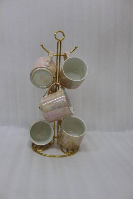 A set of 6 iridescent ceramic tea and coffee mugs hanging on a gold-tone metal stand resting on a wooden tray with a kitchen interior background.