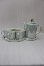 Luxurious iridescent ceramic tea set featuring six cups, one kettle, and a gold-finish stand on a white tray, arranged on a marble kitchen counter.