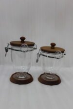 Set of three clear acrylic jars of varying sizes with wooden bases, wooden lids, airtight metal clamps, and hanging acrylic spoons on a kitchen countertop.