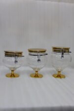 Three graduated luxury clear acrylic storage jars on pedestals with gold bases, matching gold lids, silver locking latches, and spoons arranged on a marble countertop.