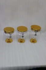 Three graduated luxury clear acrylic storage jars on pedestals with gold bases, matching gold lids, silver locking latches, and spoons arranged on a marble countertop.
