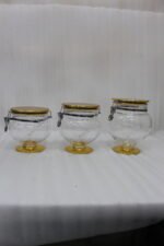 Three graduated clear acrylic jars with gold bases, lids, and matching spoons displayed on a dark countertop from One More Crockery.