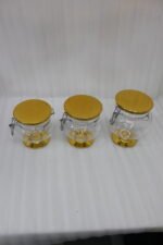 Three graduated clear acrylic jars with gold bases, lids, and matching spoons displayed on a dark countertop from One More Crockery.
