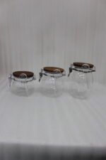 Three clear acrylic storage jars of varying sizes featuring wooden lids, airtight wire-clamp seals, and small hanging spoons, arranged on a white marble countertop in a modern kitchen.