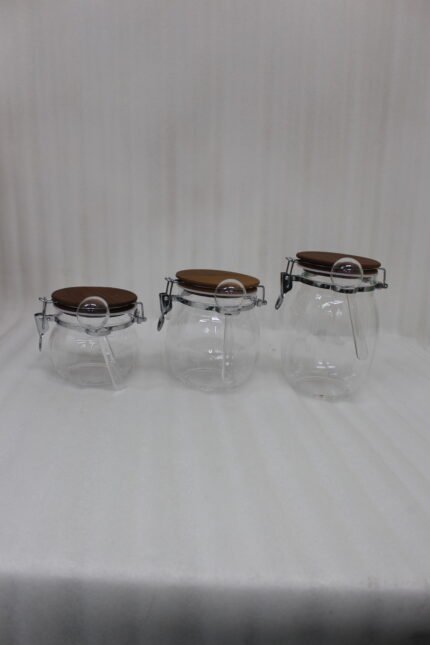 Three clear acrylic storage jars of varying sizes featuring wooden lids, airtight wire-clamp seals, and small hanging spoons, arranged on a white marble countertop in a modern kitchen.