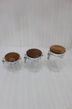 Clear acrylic kitchen storage jar set of three in varying sizes with wooden lids, airtight clasp locks, and attached spoons sitting on a dark marble countertop against a modern kitchen background.
