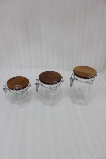 Clear acrylic kitchen storage jar set of three in varying sizes with wooden lids, airtight clasp locks, and attached spoons sitting on a dark marble countertop against a modern kitchen background.